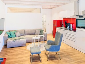 Ein modernes Wohnzimmer mit einer Couch, zwei Sesseln, einem Couchtisch und einer Küchenzeile mit einem roten Kühlschrank und einem Herd.