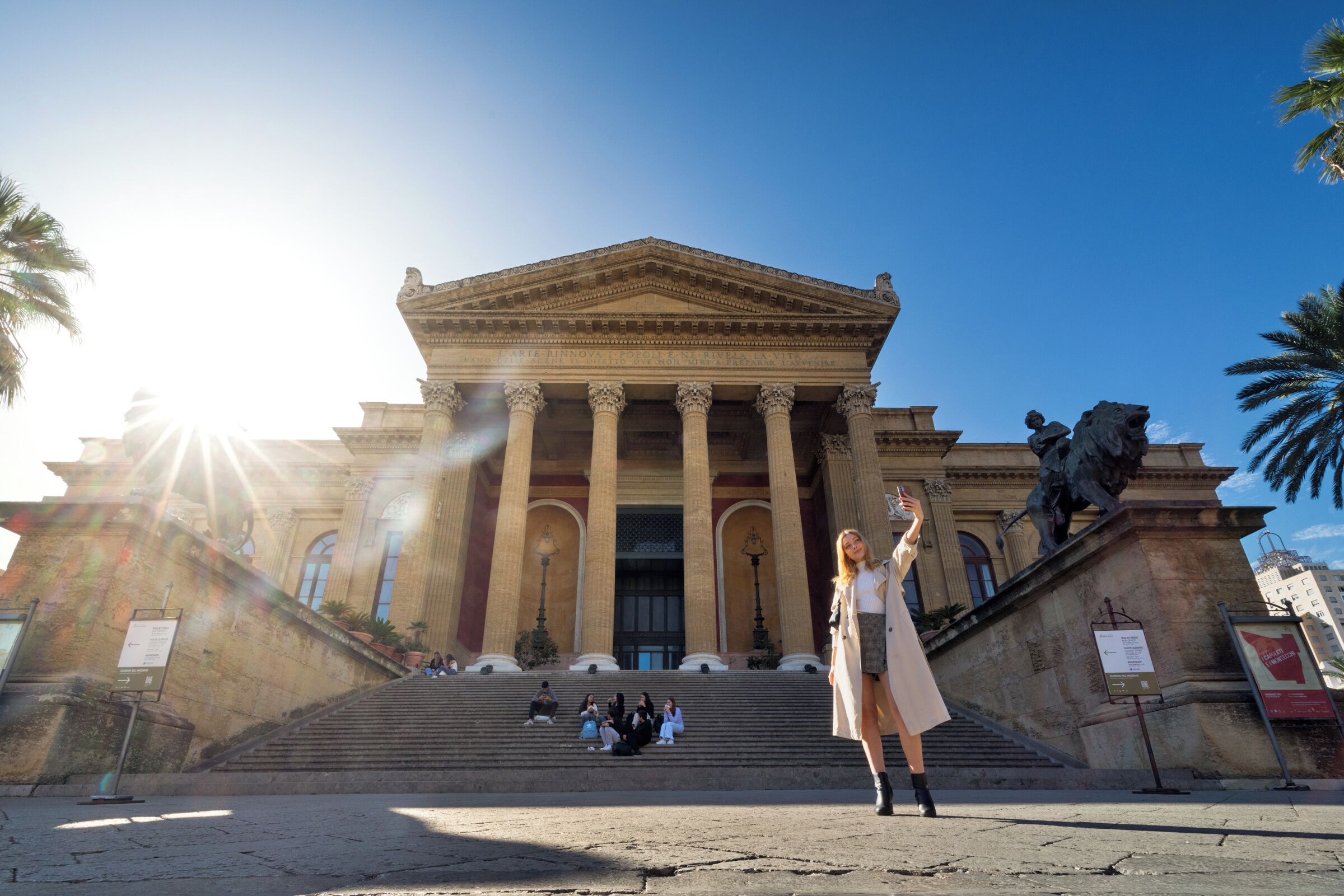A grand building with classical architecture, featuring a large staircase leading to the entrance, palm trees, and a statue on the right.