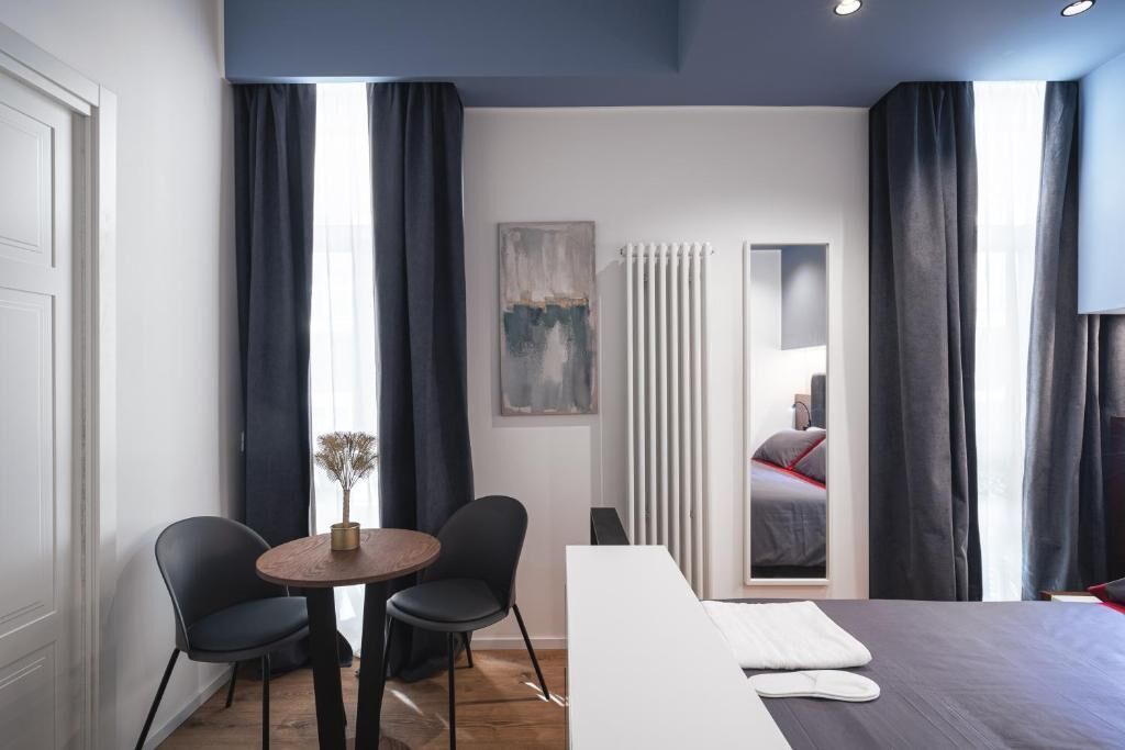 A modern hotel room with a large bed, a dining table with chairs, and a view of another room through a mirror.