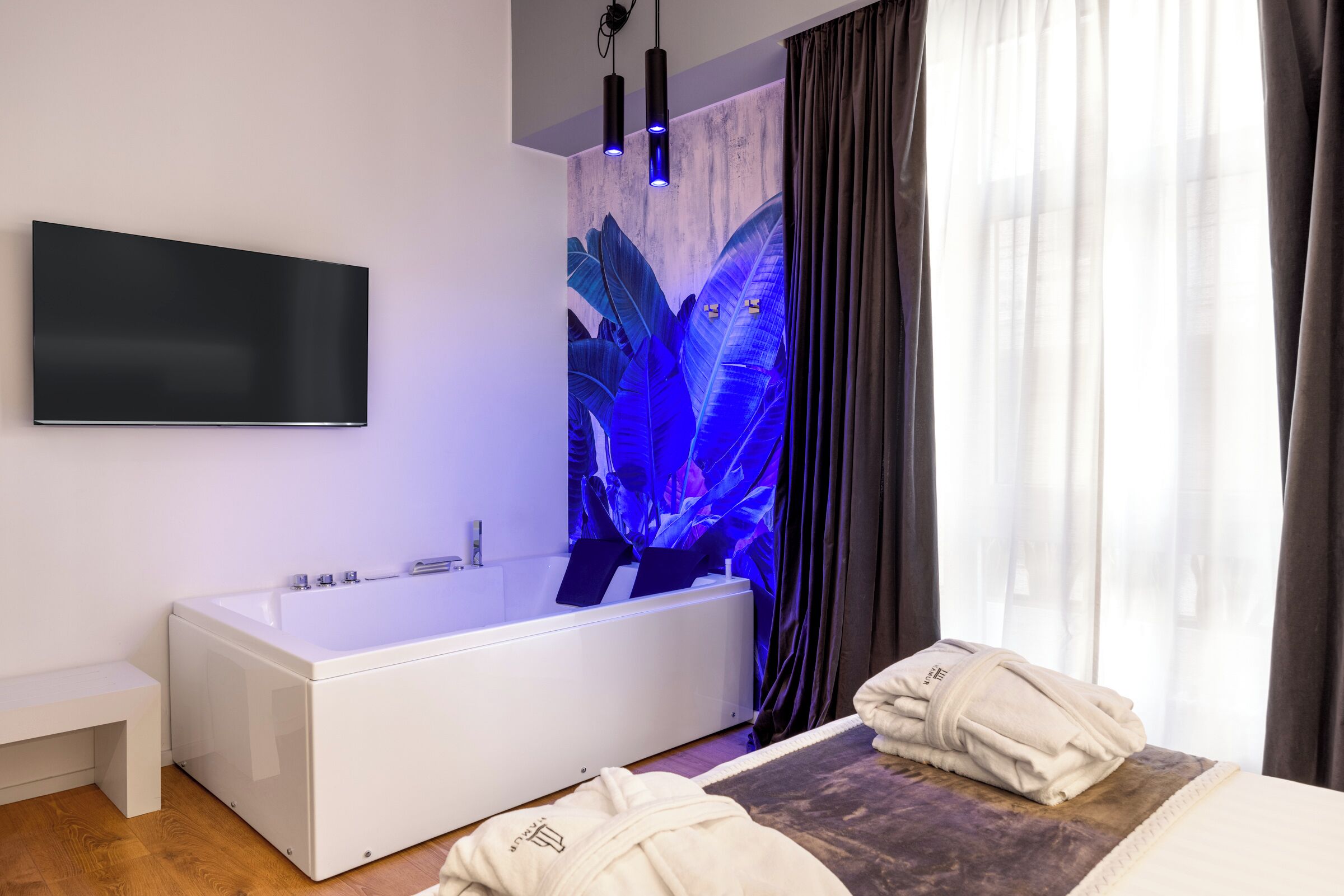 Une salle de bain moderne dotée d’une grande baignoire, d’un téléviseur à écran plat fixé au mur et d’une grande fenêtre avec des rideaux.