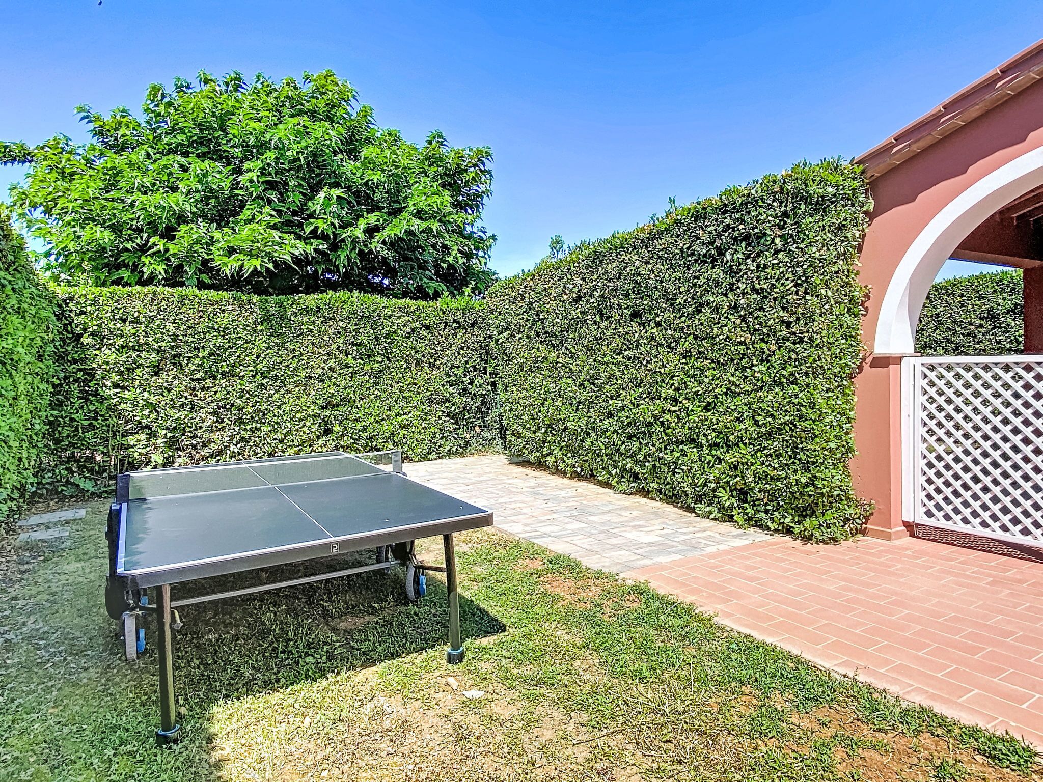 A ping pong table in a garden with a hedge and a brick wall.