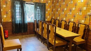 A dining room with a long wooden table and chairs, a fire extinguisher, and a window with curtains. - Lakshmi Guest House (Srinagar)