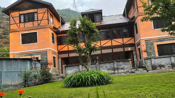 A two-story building with a stone foundation and wooden upper structure, surrounded by a well-maintained lawn with orange flowers. - Lakshmi Guest House (Srinagar)