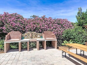 Un espace barbecue extérieur avec des barbecues en briques, un banc en bois et un décor composé d’arbustes en fleurs.