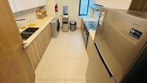 A compact kitchen with a stainless steel refrigerator, a washing machine, a water dispenser, and a trash bin. - emaro hostel (Dubai)