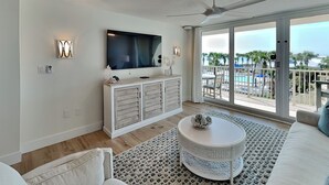 Ein modernes Wohnzimmer mit einem Flachbildfernseher, einem Deckenventilator, einem weißen Sofa, einem runden Couchtisch und einem Balkon mit Blick auf einen Pool.
