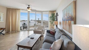 A spacious living room with a large window, a ceiling fan, a sofa, a coffee table, and a painting on the wall. - Caribe Resort 309D by Liquid Life (Orange Beach)
