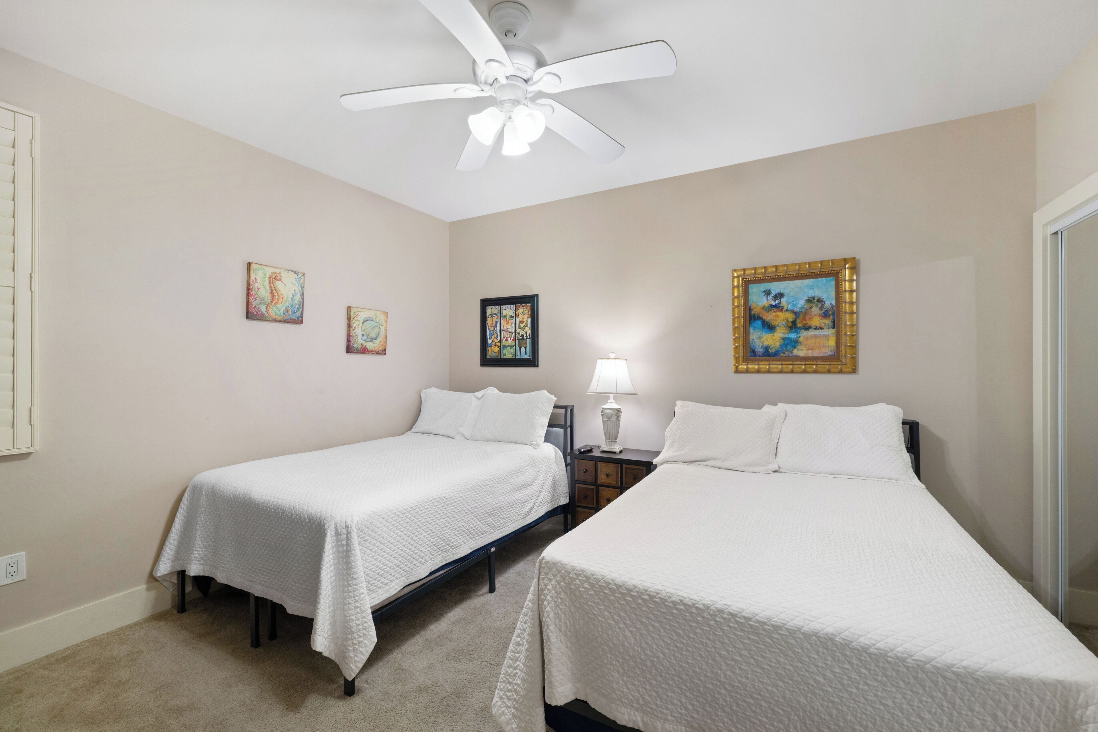 A hotel room with two beds, a ceiling fan, a nightstand, and artwork on the walls.