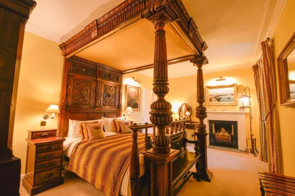 A four-poster bed with a wooden canopy, a fireplace, and a mirror on the wall.