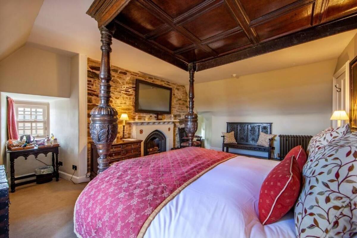 A four-poster bed with a stone wall and fireplace, a television mounted above it, and a window with curtains.
