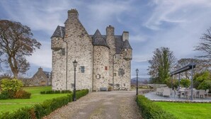 A stone castle with a paved driveway leading to a seating area with tables and chairs. - Barcaldine Castle (Oban)