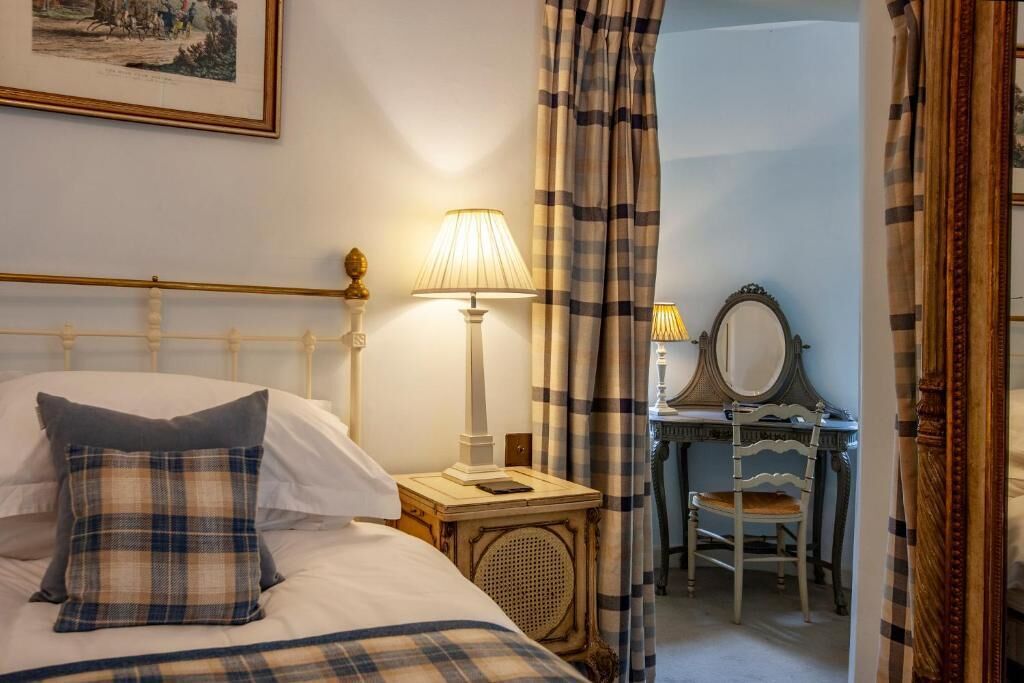 A bedroom with a bed, a bedside table with a lamp, a chair, a desk with a mirror, and curtains.
