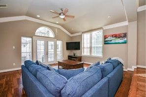 A spacious living room with a large blue sectional sofa, a wooden coffee table, a ceiling fan, and a TV mounted on the wall. - Fully Furnished 3BR / 3BA Home Near Medical Center (Houston)