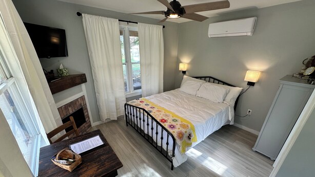 A bedroom with a bed, bedside lamps, a ceiling fan, a TV, a desk with a chair, and a window with curtains.