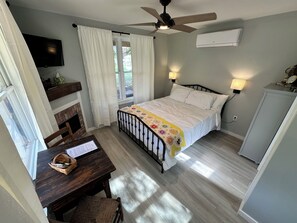 A bedroom with a bed, bedside lamps, a ceiling fan, a TV, a desk with a chair, and a window with curtains.