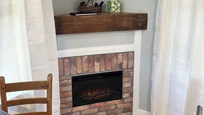 A room with a flat-screen TV mounted above a fireplace, a wooden table with a chair, and a wicker chair.