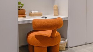 Un baño moderno con un tocador blanco, un espejo, un televisor montado sobre el espejo, una planta en maceta y una silla naranja original.