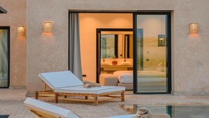 Un edificio moderno con dos sillones de descanso en el exterior, una puerta de cristal que da a una habitación con cama y baño a la vista.