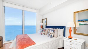 Een slaapkamer met een groot raam, een bed met een blauwe hoofdbord, een witte dressoir en een spiegel aan de muur.