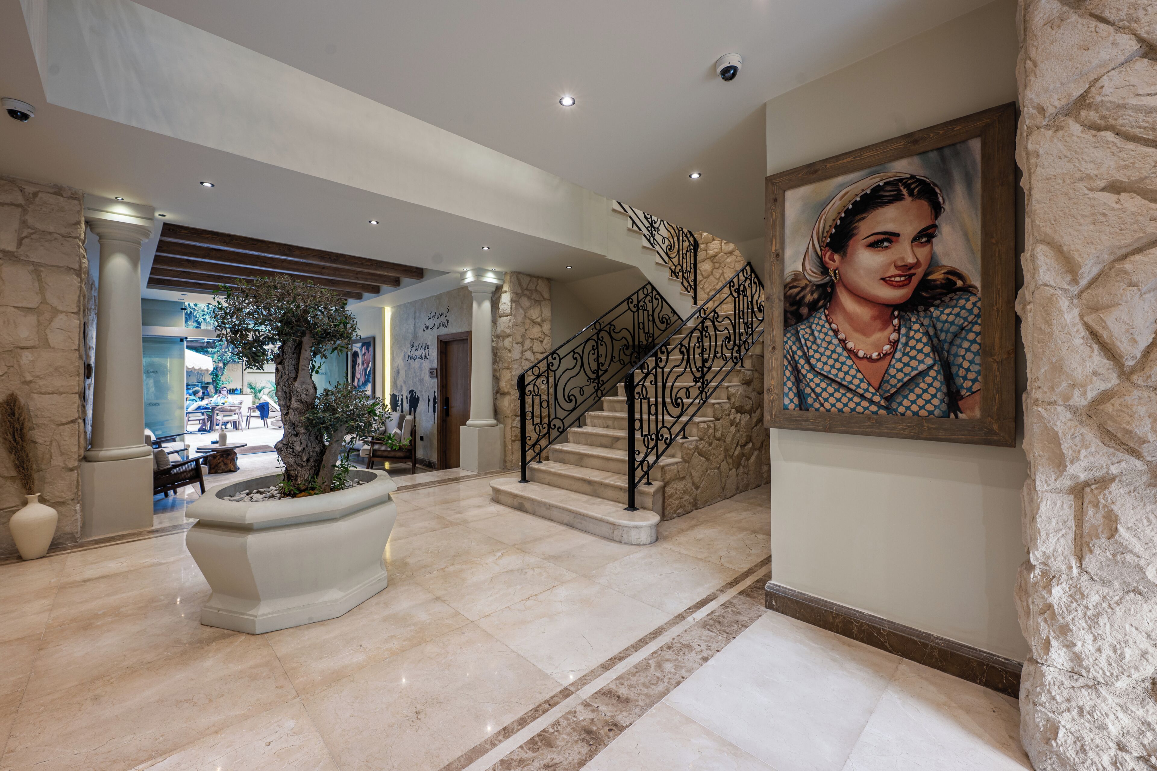 Un hall d’entrée avec un sol en marbre, un escalier orné de balustrades travaillées, un grand arbre en pot et un portrait encadré accroché au mur.