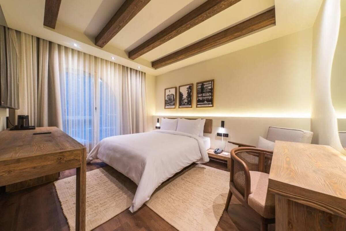 A bedroom with a bed, bedside tables, a wooden desk, a chair, and a large window with curtains.