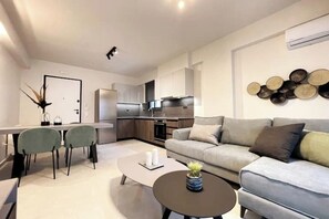 A modern living room with a sofa, coffee table, and chairs. There is a kitchen area with cabinets and appliances in the background. - Athenean Gem Near Athens Metro Mall-B2 (Ilioupoli)