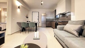 A modern living room with a sofa, a coffee table, and a dining area with chairs. There is a kitchen with a refrigerator and stove in the background. - Athenean Gem Near Athens Metro Mall-B2 (Ilioupoli)