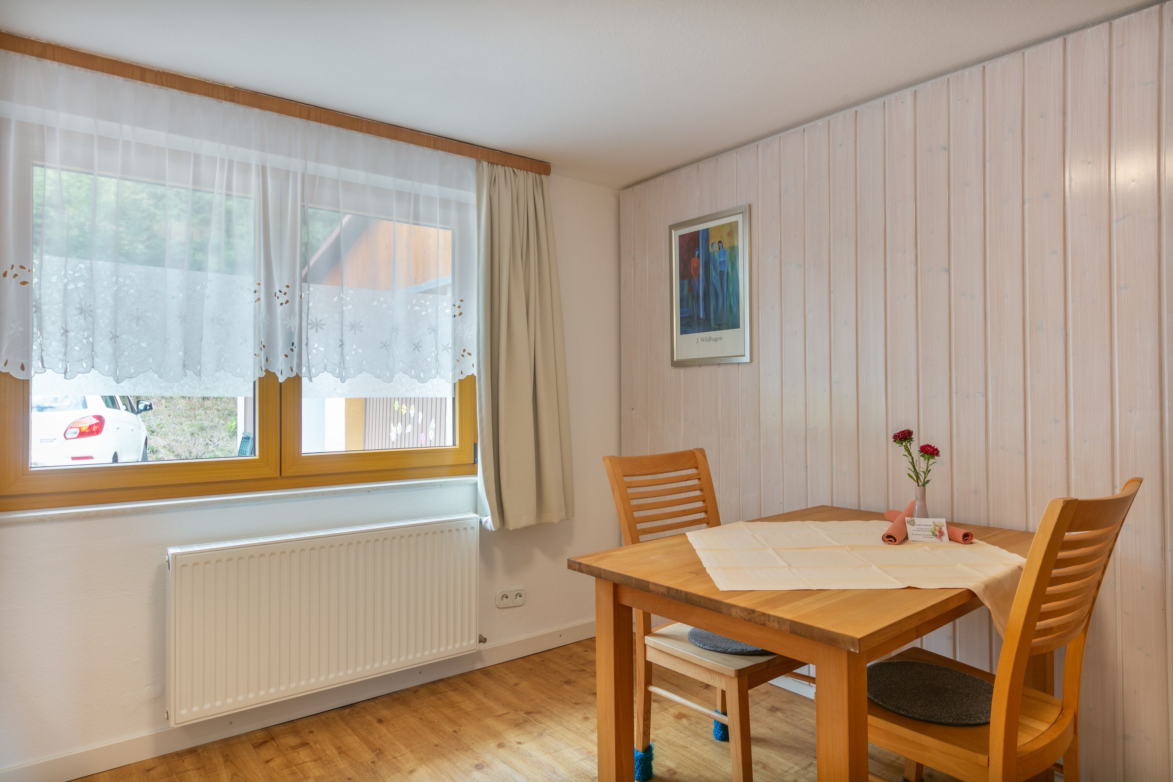 A room with a wooden table, chairs, a radiator, a window with curtains, and a framed painting on the wall.