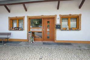 Un edificio con puerta de madera, dos ventanas con contraventanas, un banco y un pequeño jardín.