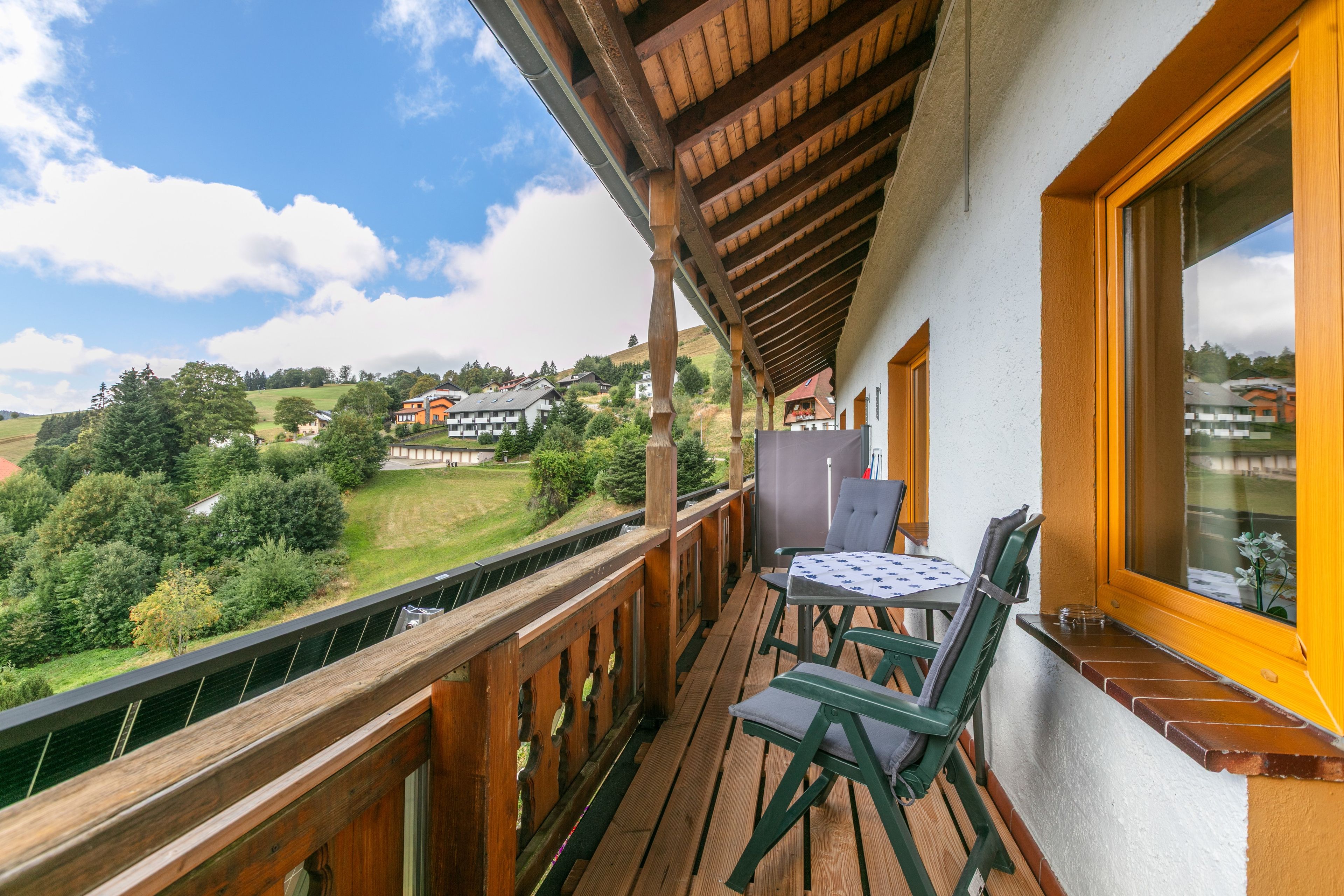 A balcony with a wooden railing, a table with chairs, and a window overlooking a hillside with buildings.