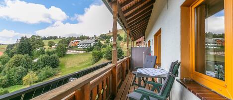 A balcony with a wooden railing, a table with chairs, and a window overlooking a hillside with buildings.