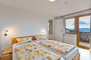 A double bed with floral bedding, a bedside table, a wall-mounted lamp, and a view of the outdoors through a sliding door. - Alpenblick Apartment (Todtnau)