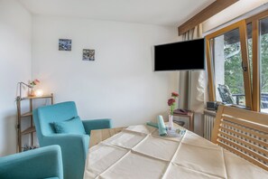Une pièce avec un canapé de couleur turquoise, une table recouverte d’une nappe, un téléviseur à écran plat et une fenêtre avec des rideaux.
