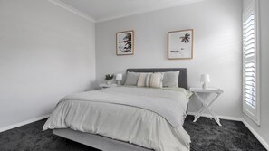 Une chambre à coucher comprenant un lit, deux tables de chevet, une commode, une fenêtre avec des stores et deux tableaux encadrés accrochés au mur.