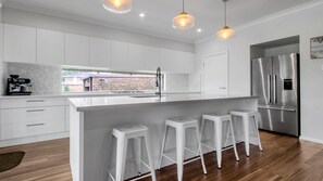 A modern kitchen with a large island, bar stools, stainless steel appliances, and a view of a brick building. - Mibilla House in Sussex Inlet (Sussex Inlet)
