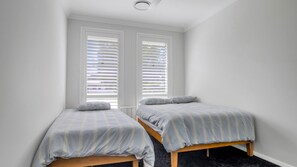 A bedroom with two single beds, white walls, and a window with blinds. - Mibilla House in Sussex Inlet (Sussex Inlet)