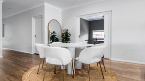 A modern dining area with a round table and four white chairs, a mirror, and two potted plants. - Mibilla House in Sussex Inlet (Sussex Inlet)