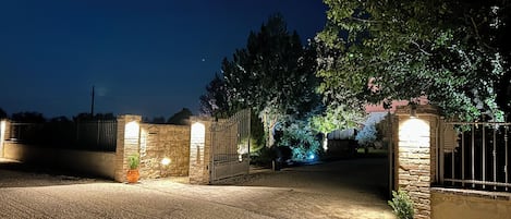 Eine Einfahrt mit Tor, nachts beleuchtet, mit Steinmauern und einem Topfpflanzen.