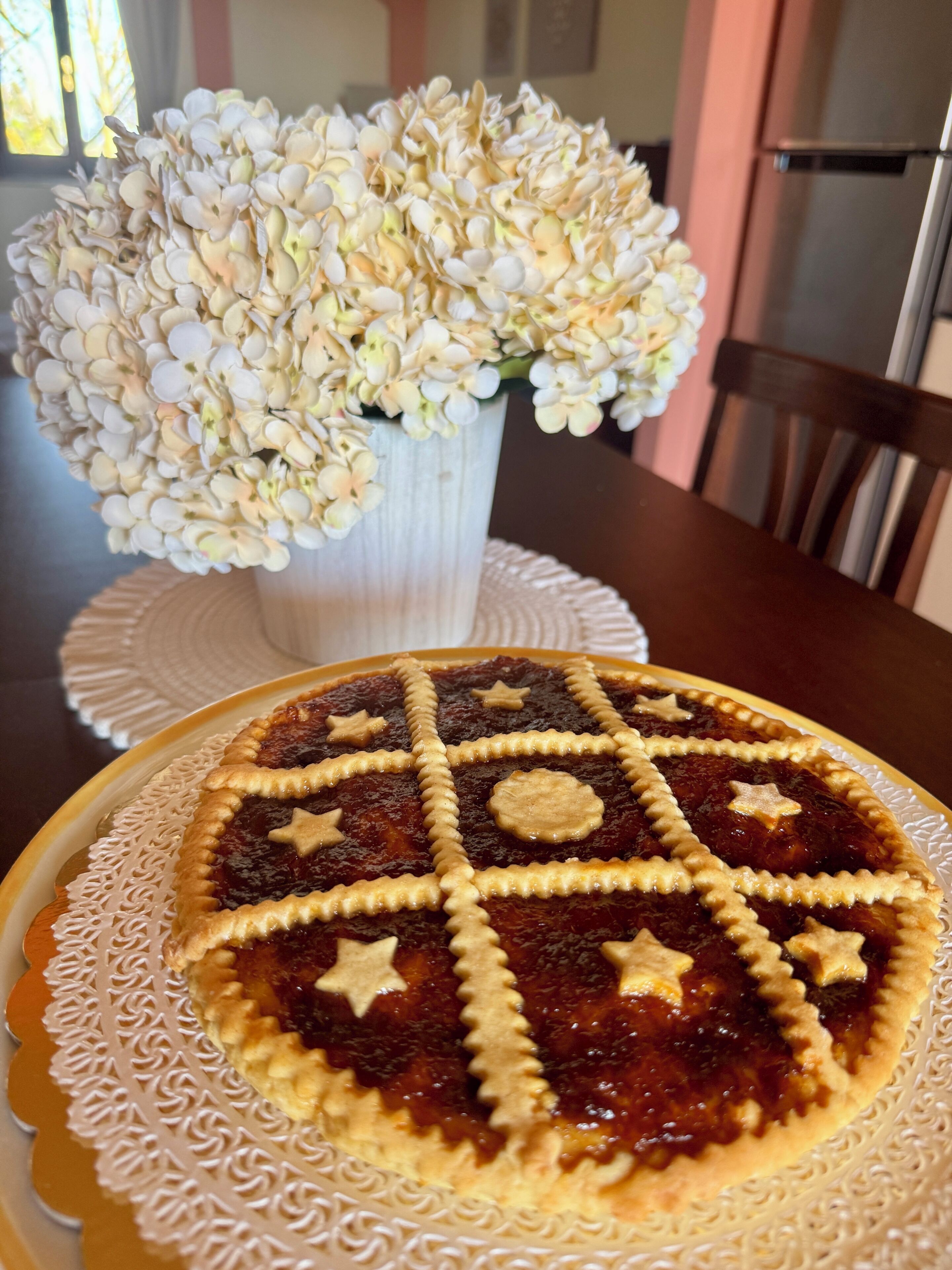 A pie with star-shaped cutouts on a decorative plate, a white vase with flowers, and a dining table.
