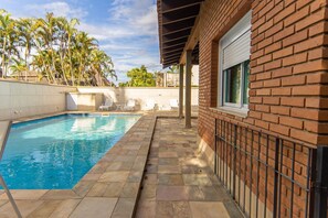 A swimming pool with a tiled deck, a brick wall, and a covered patio area. - Casa com 3 Suítes, Piscina e Churrasqueira (Guarujá)