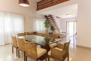 A modern living room with a glass-top dining table, wicker chairs, a sofa, a staircase, and a potted plant. - Casa com 3 Suítes, Piscina e Churrasqueira (Guarujá)