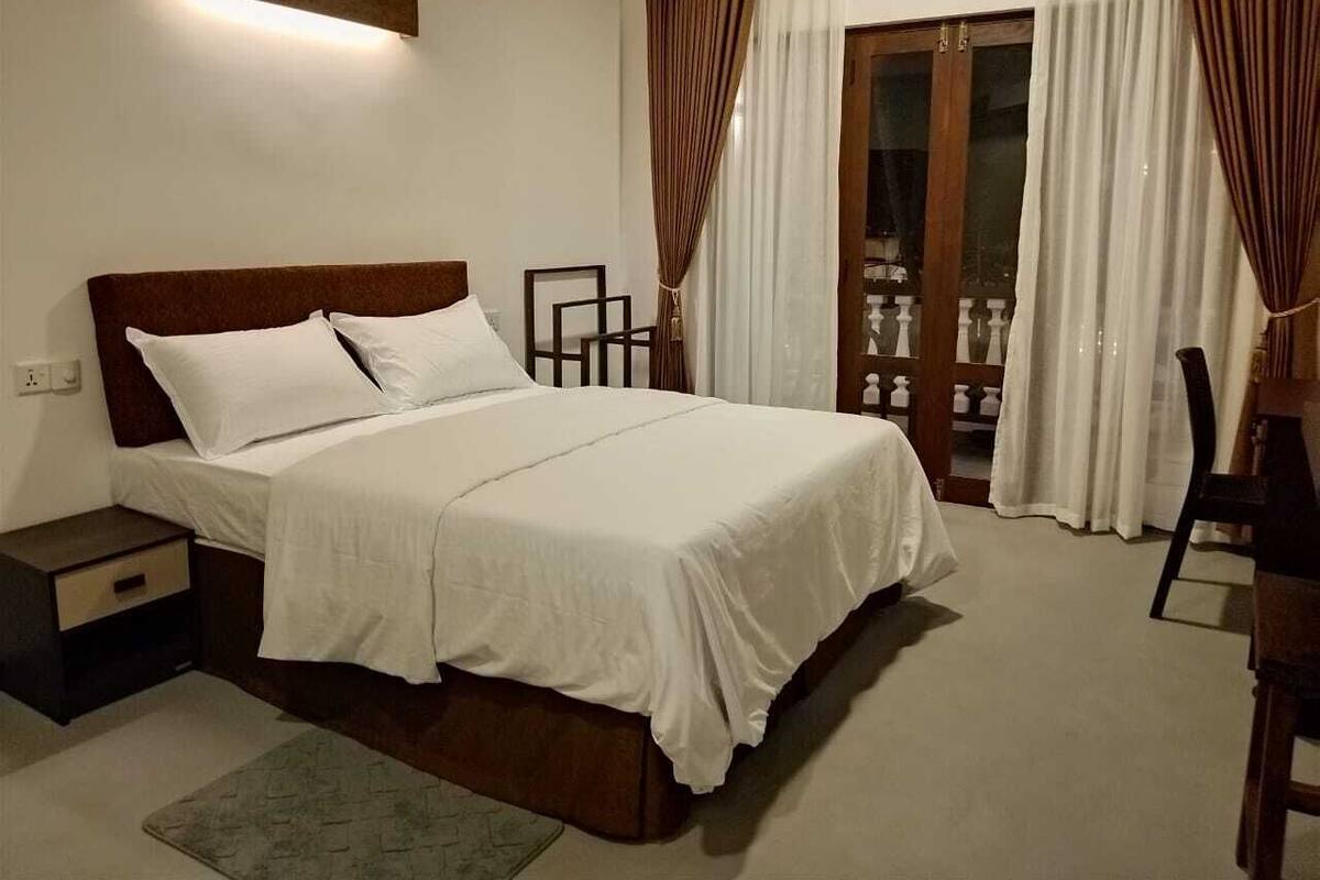 A bedroom with a bed, bedside tables, a nightstand, a chair, and a view of a balcony with curtains.