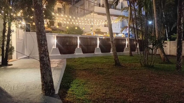 Un espacio al aire libre de noche, con luces colgantes, árboles y una cerca blanca.