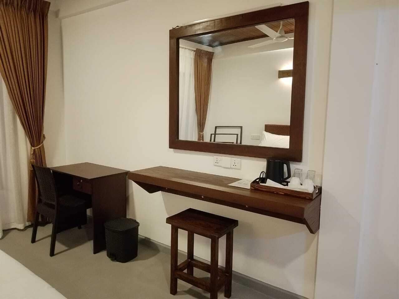 A hotel room with a wooden desk, a chair, a small stool, a trash bin, a mirror, and a coffee maker.