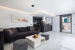 A modern living room with a sofa, coffee table, and ottoman, a kitchen in the background, and a door leading to an outdoor area.