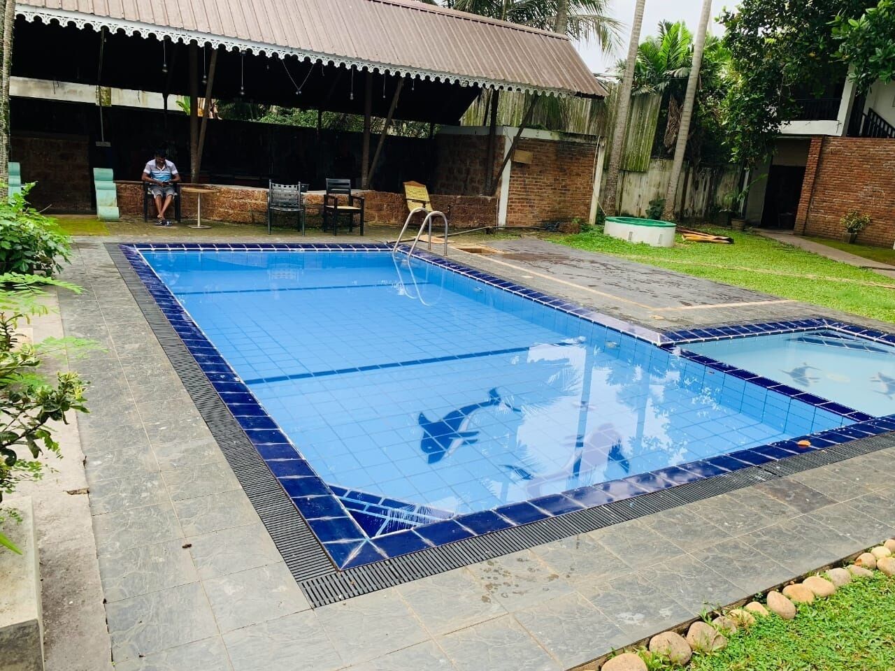 A rectangular swimming pool with a tiled edge, a covered seating area, and a brick wall in the background.
