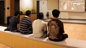 A group of people seated on a bench watching a presentation in a room with a projector screen. - C Suites Gilbert (Bengaluru)