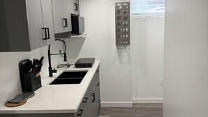 Una cocina compacta con cubierta blanca, armarios grises, un basurero negro y una ventana con persianas.