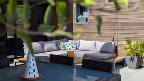 A modern outdoor seating area with a glass table, wicker chairs, and a wooden wall. - Guest Room 'L'Abri des Flots' with Shared Pool, Terrace, and Wi-Fi (Plouër-sur-Rance)