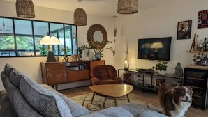A living room with a sofa, a coffee table, a TV, and a dog. - Guest Room 'L'Abri des Flots' with Shared Pool, Terrace, and Wi-Fi (Plouër-sur-Rance)